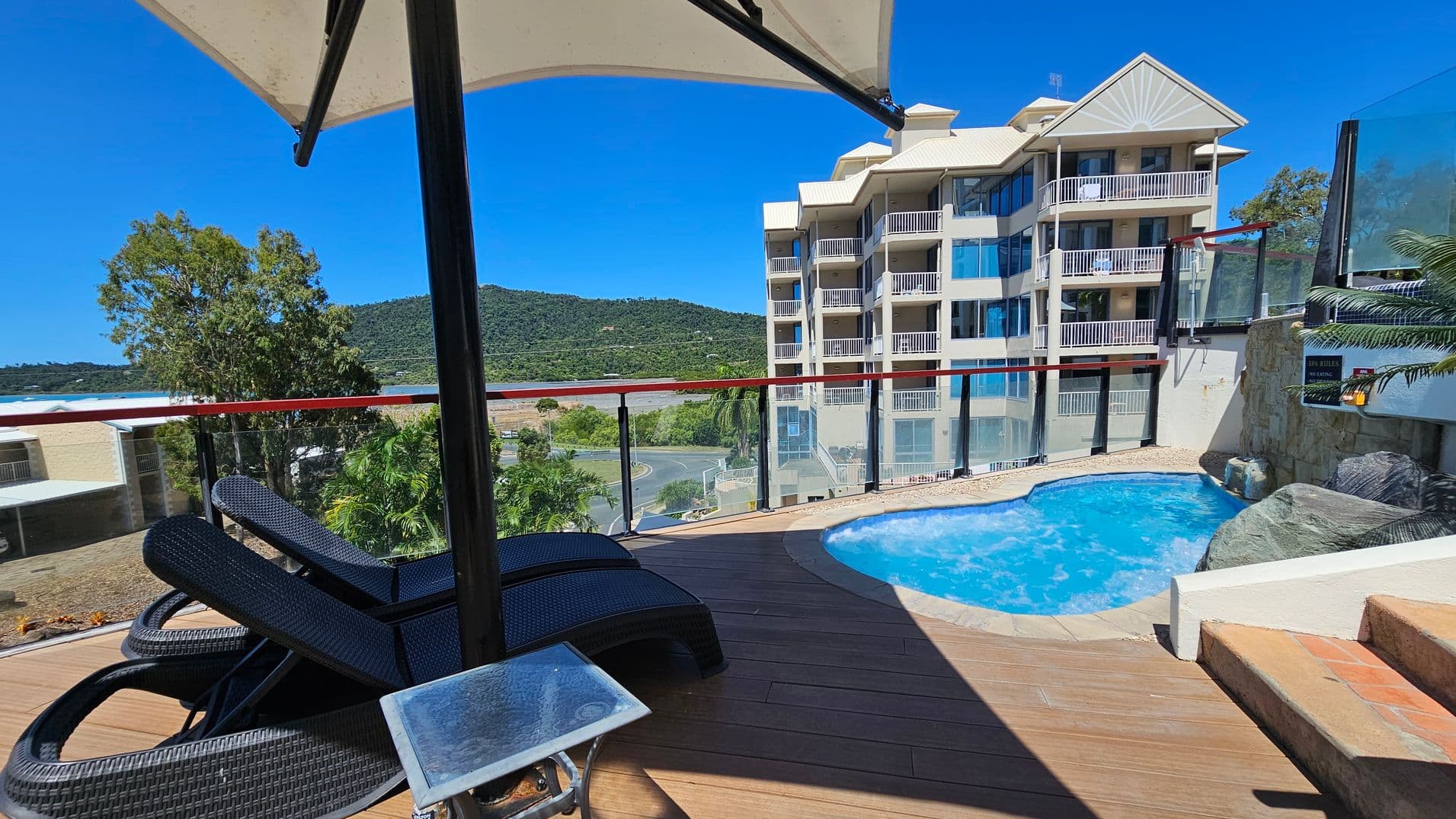 A poolside area with two black wicker sun loungers on the left, shaded by a large umbrella. A small glass-topped table is nearby. The pool is on the right, surrounded by a stone deck. In the background, a multi-storey building with balconies is visible, along with a view of green hills and trees. A glass railing runs along the edge of the deck, providing a clear view of the landscape.