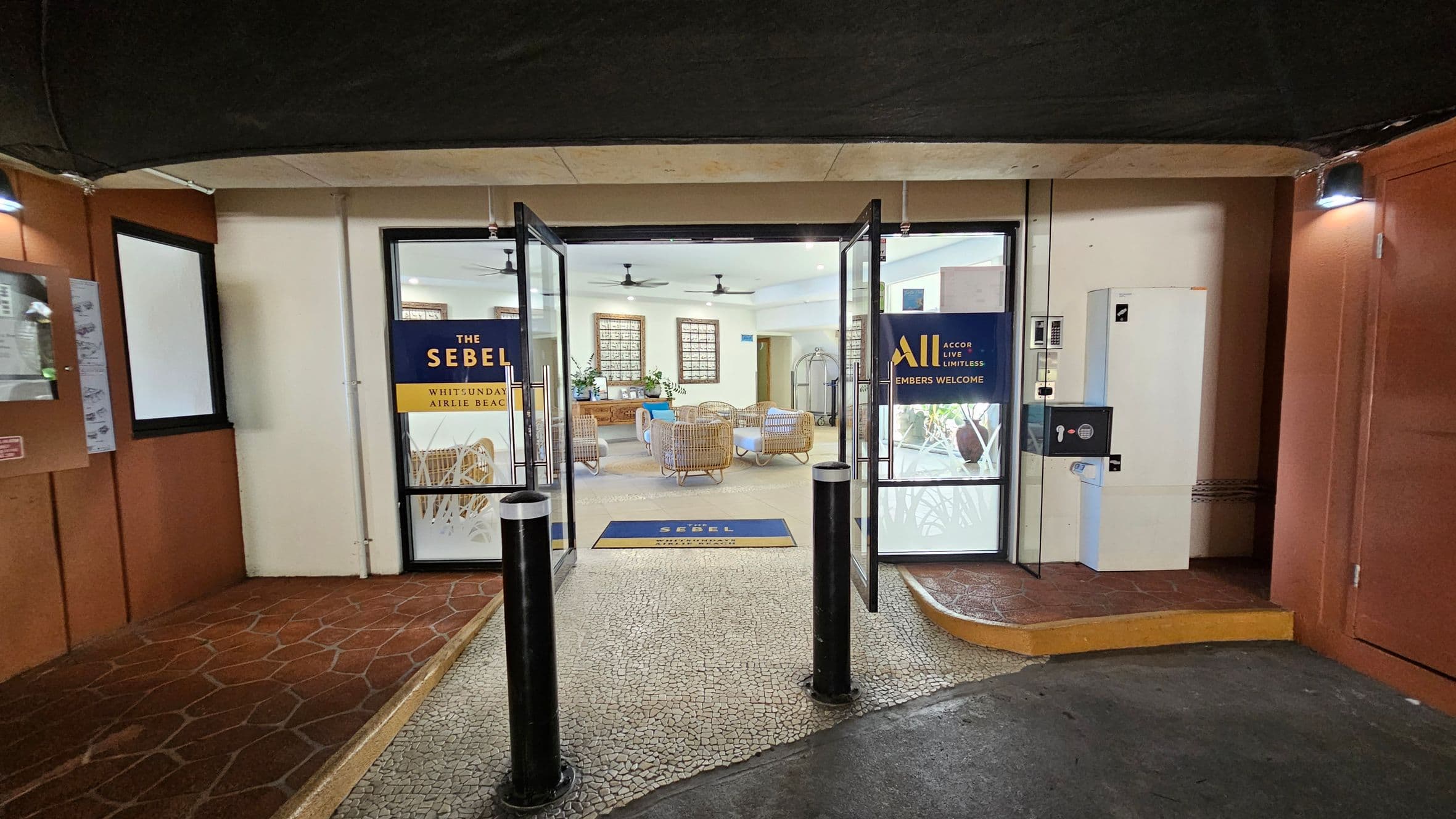 Glass doors lead into a hotel lobby with wicker furniture and ceiling fans. Signs on the doors read "The Sebel Whitsunday Airlie Beach" and "Accor Live Limitless Members Welcome." The floor outside is a mosaic pattern, transitioning to a tiled floor inside. On the left, a bulletin board is mounted on a brown wall. On the right, a white utility box and a keypad are attached to the wall. The ceiling is dark, contrasting with the lighter interior.