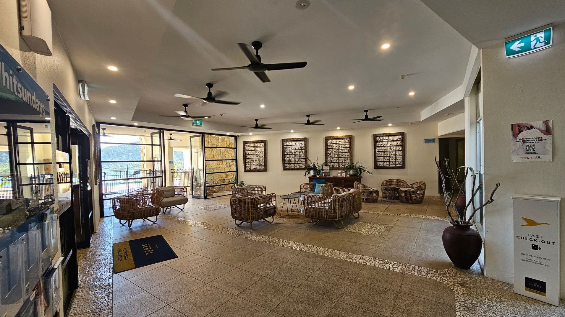 A spacious lobby area with wicker seating arranged around small tables in the centre. The floor is tiled with a mosaic pattern. On the left, glass doors lead to an outdoor area with a view of hills and water. The ceiling has multiple ceiling fans and recessed lighting. On the right, a large vase with branches and a sign for fast check-out are visible. The back wall features framed artwork and a wooden console table with plants.