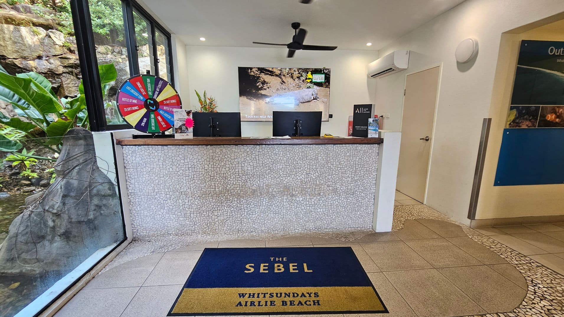 Reception area with a mosaic-tiled counter in the centre, featuring two computer monitors. A colourful prize wheel is on the left side of the counter. Behind the counter, a large screen displays an image of a crocodile. To the right, a door and a wall-mounted air conditioning unit are visible. A blue and gold mat on the floor reads "The Sebel Whitsundays Airlie Beach." Large windows on the left reveal lush greenery and rocks outside. The ceiling has recessed lighting and a black ceiling fan.