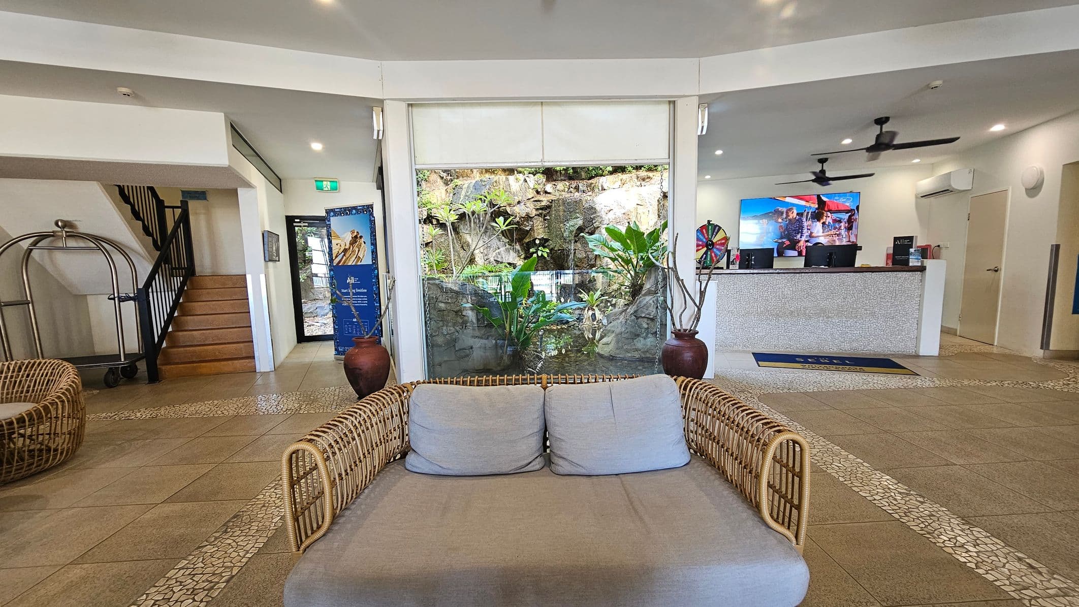 A hotel lobby with a wicker sofa in the foreground. In the centre, a large window displays a rock garden with lush green plants. To the left, a staircase with a luggage cart nearby. On the right, a reception desk with a colourful display screen and a spinning wheel. The floor is tiled with a mosaic pattern, and the ceiling has recessed lighting and ceiling fans.