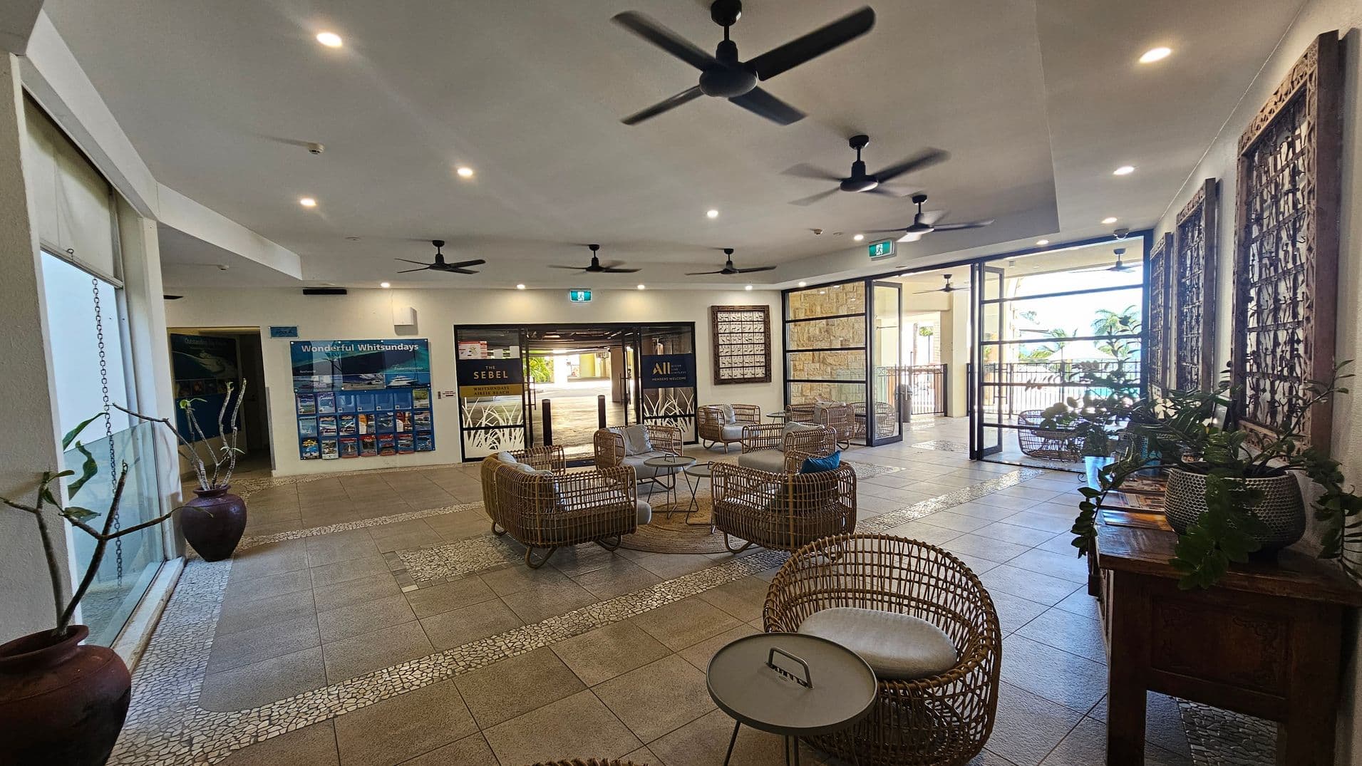 Spacious lobby area with wicker seating arranged in a circular pattern around small tables. The floor is tiled with a mosaic design. On the left, large windows with potted plants. The back wall features travel brochures and a sign for "Sebel." The right side has decorative wooden panels and more plants on a wooden table. Ceiling fans and recessed lighting are visible above. The entrance has glass doors leading to an outdoor area with palm trees.