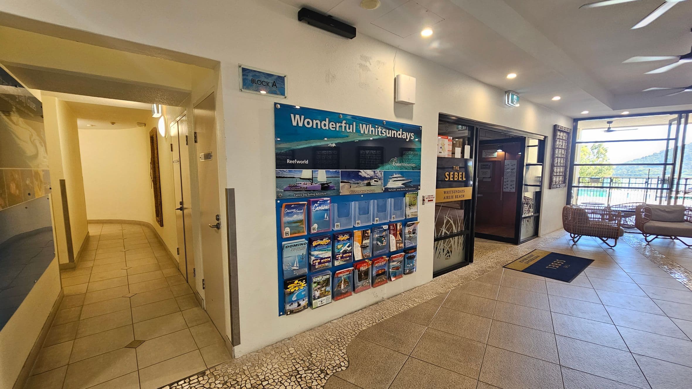 A hallway with tiled flooring leads to the left, with doors on the right side. On the right wall, a display board titled "Wonderful Whitsundays" features brochures and information. Next to it, a glass door with "The Sebel" signage opens to a room. Beyond, large windows reveal a view of trees and hills. The floor has a mix of mosaic and large tiles, and the ceiling is fitted with recessed lights and ceiling fans. Wicker chairs are near the windows.