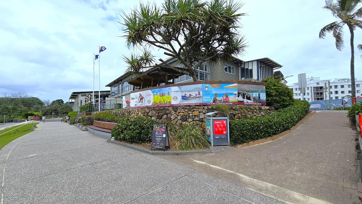 A paved pathway leads to a two-storey building with a balcony adorned with colourful banners. A tropical tree stands tall beside it. Flags on poles flank the building, with two red structures resembling phone booths or information stands nearby. A blackboard sign is on the left. The sky is overcast, with buildings in the background. Bicycle racks are visible.