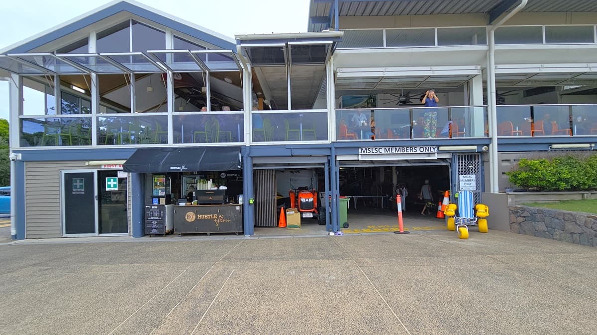 Front view of a two-storey facility building with a spacious concourse and glass railings. A drink retail shop is on the left next to the first aid room. Beside it, a tractor and an entrance sign read "MSLSC Members Only," with an orange bollard. The upper floor has arranged seating chairs. To the right is a grass area with landscaped plants.