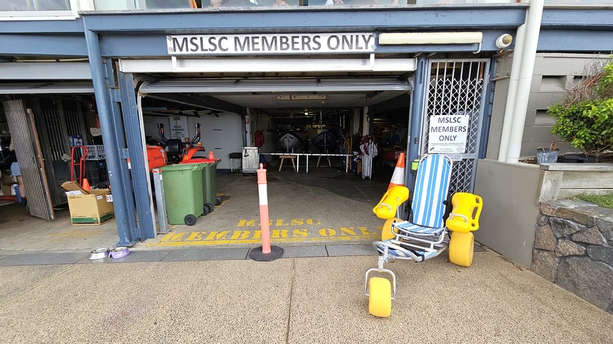 Entrance for members. On the left, a tractor; on the right, an entrance with a sign "MSLSC Members Only," marked by an orange bollard. A beach wheelchair is positioned nearby. To the right, is a grass area with landscaped plants. Inside, two rubbish bins are on the left.