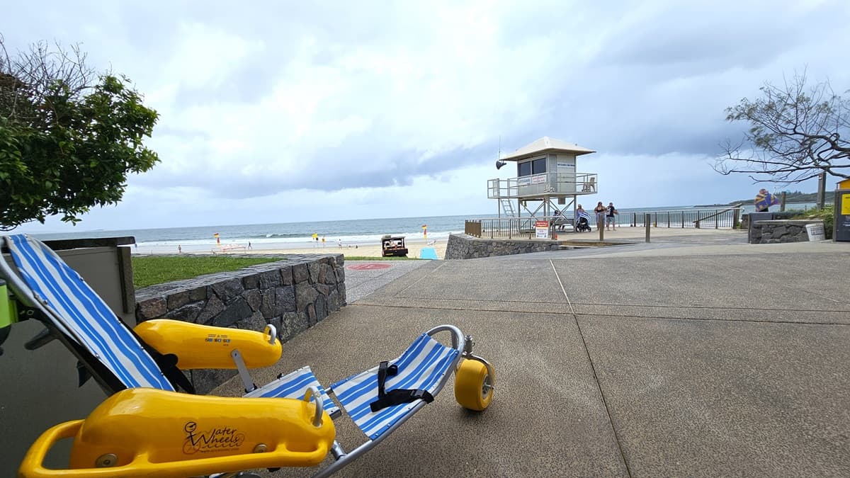A spacious concrete area facing the beach. To the left, a Mobi Chair positioned near the concrete wall. To the right, a Patrol Tower surrounded by metal railings. A landscape of grass fills the front and sides of the building.