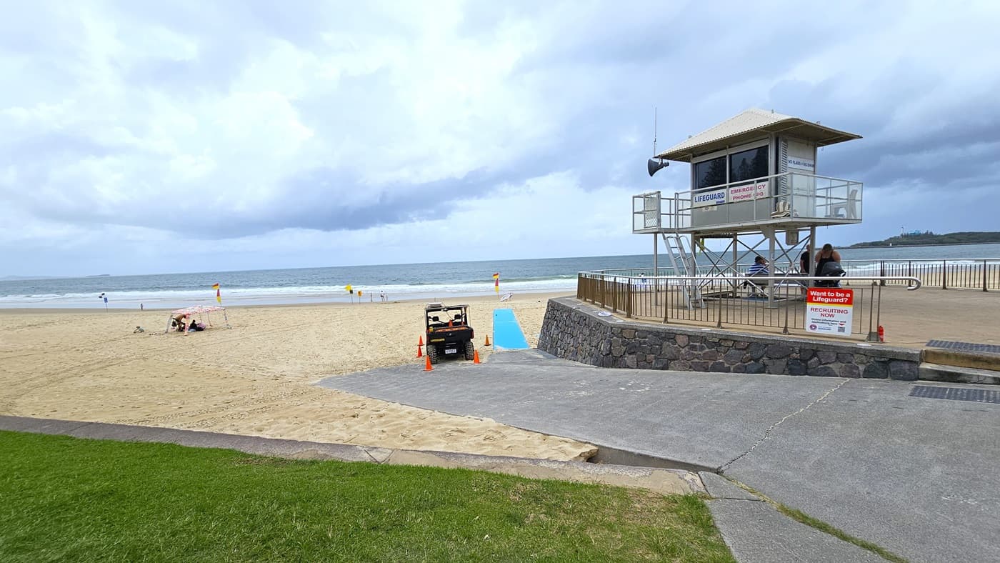 On the left, green grass lines the road. A patrol tower with stairs and railings stands on the right, surrounded by metal railings. Banners are affixed to the fence and tower. A low step with truncated domes provides access to the tower. A beach patrol car, surrounded by safety cones, is adjacent. A self-made tent is visible on the left, with the beach in the background.