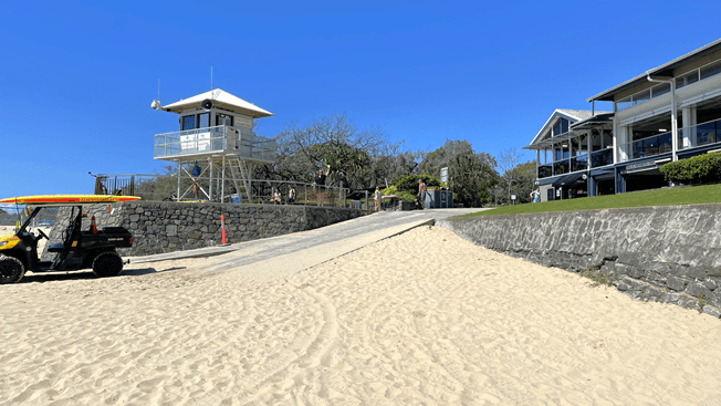 To the right, green grass lines the road. A patrol tower with stairs and railings stands on the left, surrounded by metal railings. Banners are affixed to the fence and tower. A beach patrol car is adjacent. Trees form the backdrop on the left, and a two-storey building is visible on the right.