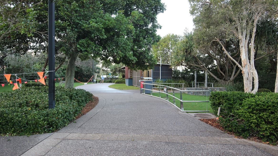 A curvy concrete pathway with a metal railing bordering the road on the right. Trimmed grass lines the roadside. To the left, a string of orange flags lines the road alongside trees and shrubs. In the distance, a low-rise structure with corrugated walls is visible.