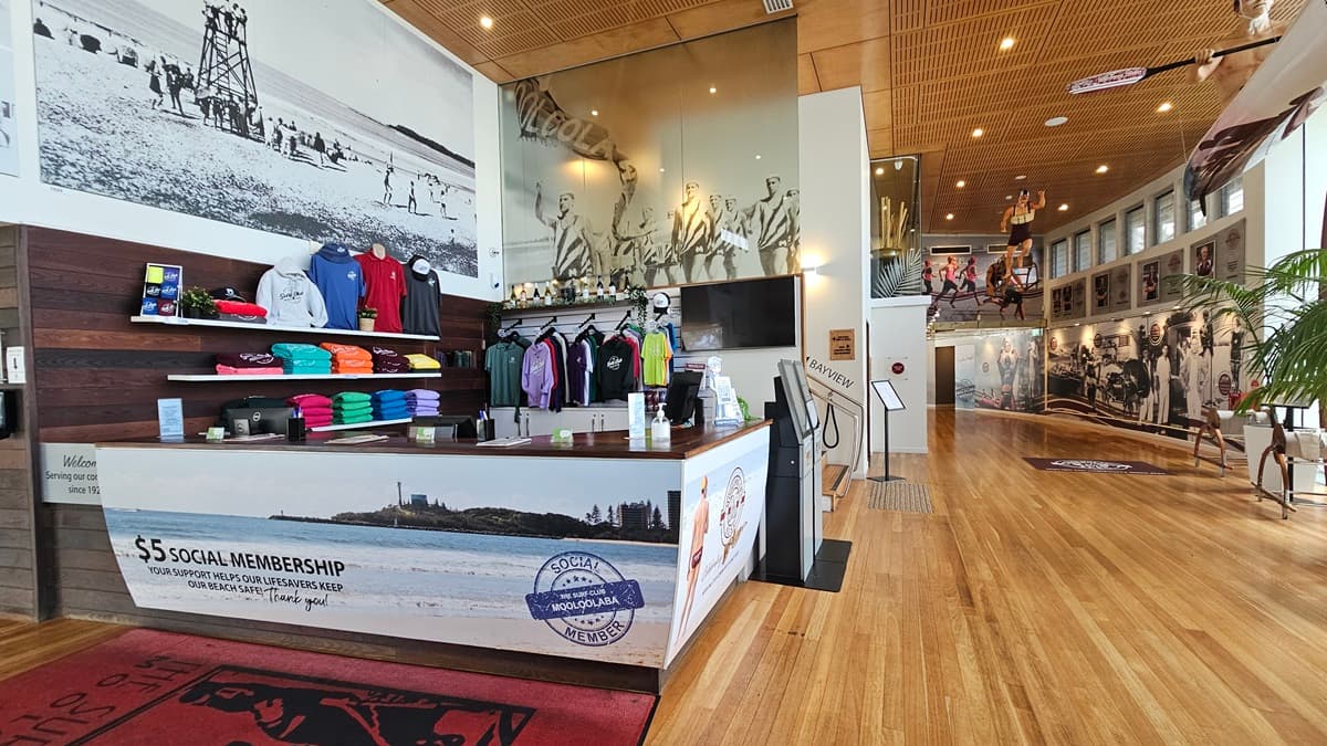 The entrance features an automatic glass sliding door equipped with a non-slip mat. A sign reading "Welcome. Enjoy your Visit!" is placed overhead. Posters are affixed to the glass door. Inside, the interior features a wooden wall with posters and timber flooring. A bench chair is positioned before the entrance, along with an automatic wet umbrella wrapper.