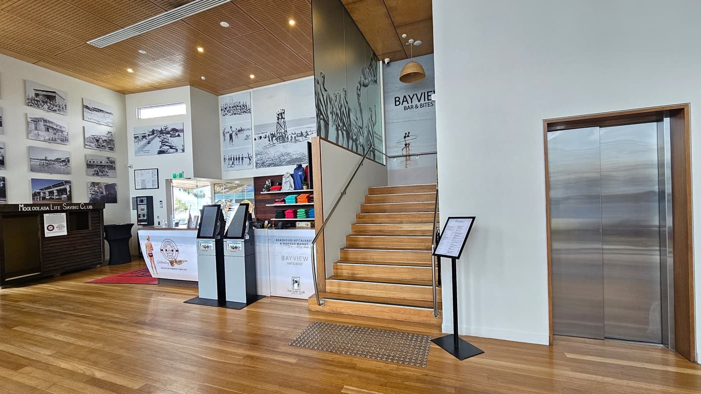 To the left is a small retail shop displaying T-shirts and jackets on a wooden display rack. Digital machines stand in front. To the right is a staircase equipped with handrails and truncated domes. A free-standing banner is positioned on the right. Adjacent is a lift. The interior features a high ceiling with large posters and imagery decorating the walls and ceiling. The floor has timber, and the ceiling has a wooden slat design with light fixtures. Large indoor plants are also visible.