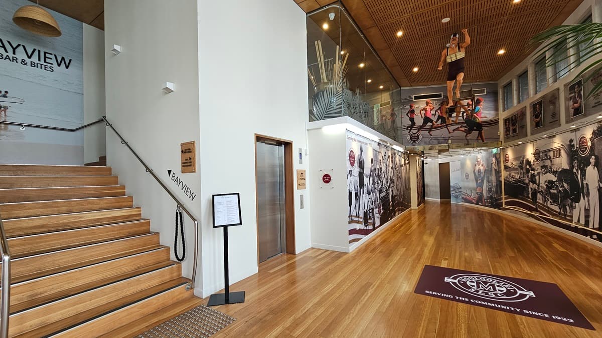 A staircase with handrails and truncated domes. A directional sign reading "Bayview" is painted on the wall alongside another poster. A free-standing banner stands on the right. Adjacent is a lift. The interior has a high ceiling adorned with large posters and imagery of a man in a surfing position. The floor is timber with a red mat featuring the facility name. The ceiling has a wooden slit design with light fixtures. Large indoor plants are visible.