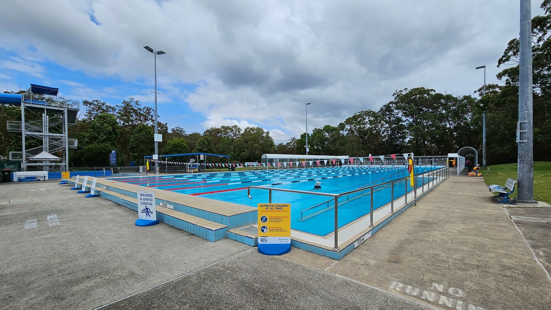 An outdoor swimming pool with marked lanes is in the centre, surrounded by a tiled deck. On the left, there's a tall water slide structure with stairs. Signs around the pool indicate safety rules. On the right, a pathway with a "No Running" warning leads to a covered area. Benches and tall lights are visible, with trees in the background. The ground is a mix of concrete and grass.