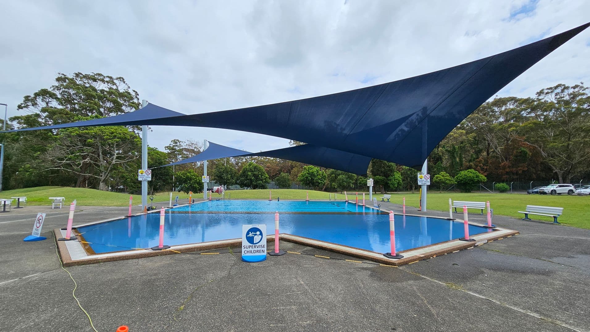 A small outdoor pool is surrounded by a paved area with a sign reading "Supervise Children" at the front. The pool is covered by large, dark blue shade sails. Pink bollards are placed around the pool's perimeter. Benches are positioned on the grass in the background, with trees and a parked car visible beyond. The ground is a mix of concrete and grass.