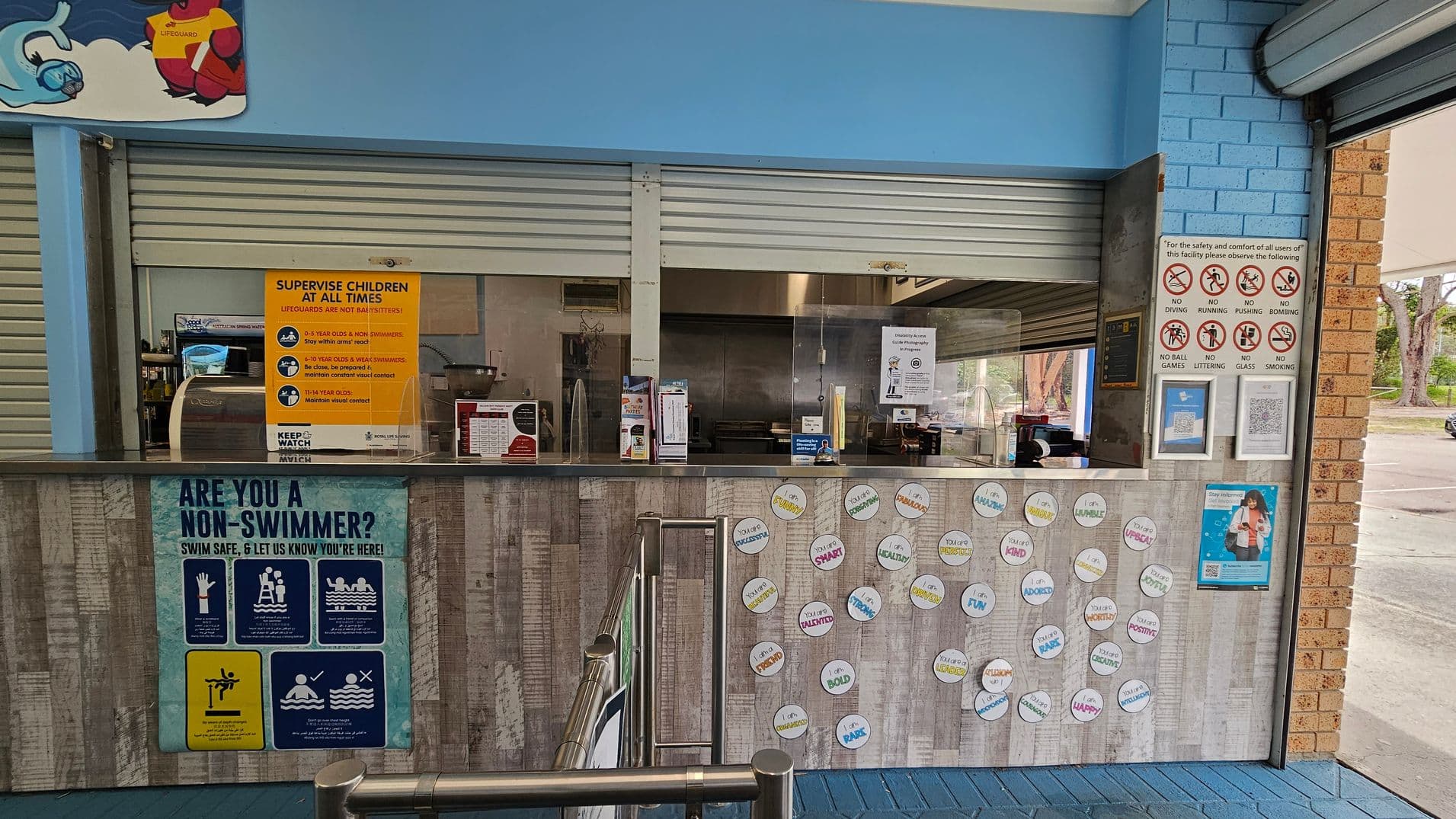 A counter area with a blue and brick wall background. The counter has various signs, including a large yellow one about supervising children and a blue one asking if you are a non-swimmer. Circular stickers with positive affirmations are displayed on the front. Behind the counter, there are closed metal shutters and a small kitchen area. On the right, there are safety signs prohibiting activities like diving and running. The floor is tiled in blue.