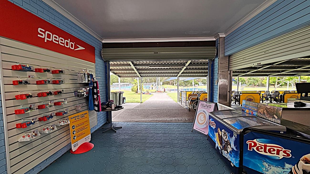 On the left, a display wall with Speedo-branded swimming accessories and a sign about supervising children. In the middle, an open area leads to an outdoor space with a path and greenery. On the right, a counter with a Peters ice cream freezer and a menu board. The floor is covered with hexagonal tiles, and the ceiling is plain.