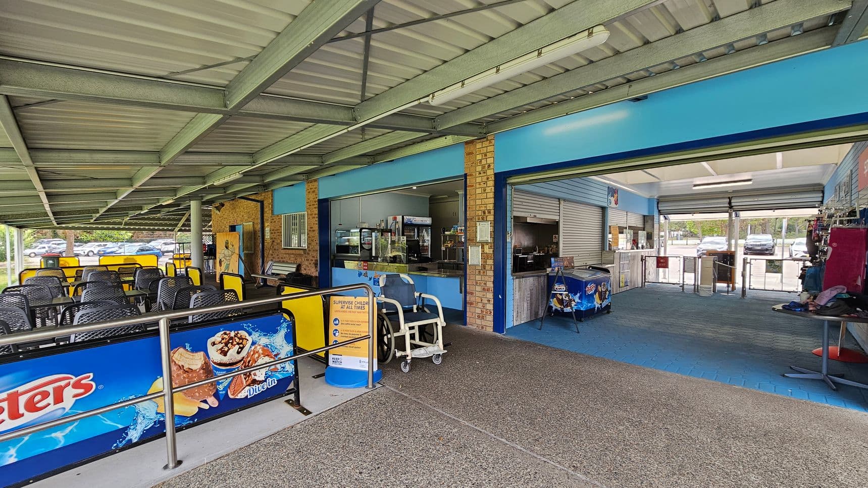 A covered outdoor seating area with black and yellow chairs on the left. The floor is a mix of textured concrete and blue tiles. In the centre, there's a counter with a display of snacks and drinks. On the right, an open area with a blue-tiled floor leads to a glass entrance with turnstiles. Ice cream advertisements are visible on barriers and a freezer. The ceiling is metal with exposed beams.
