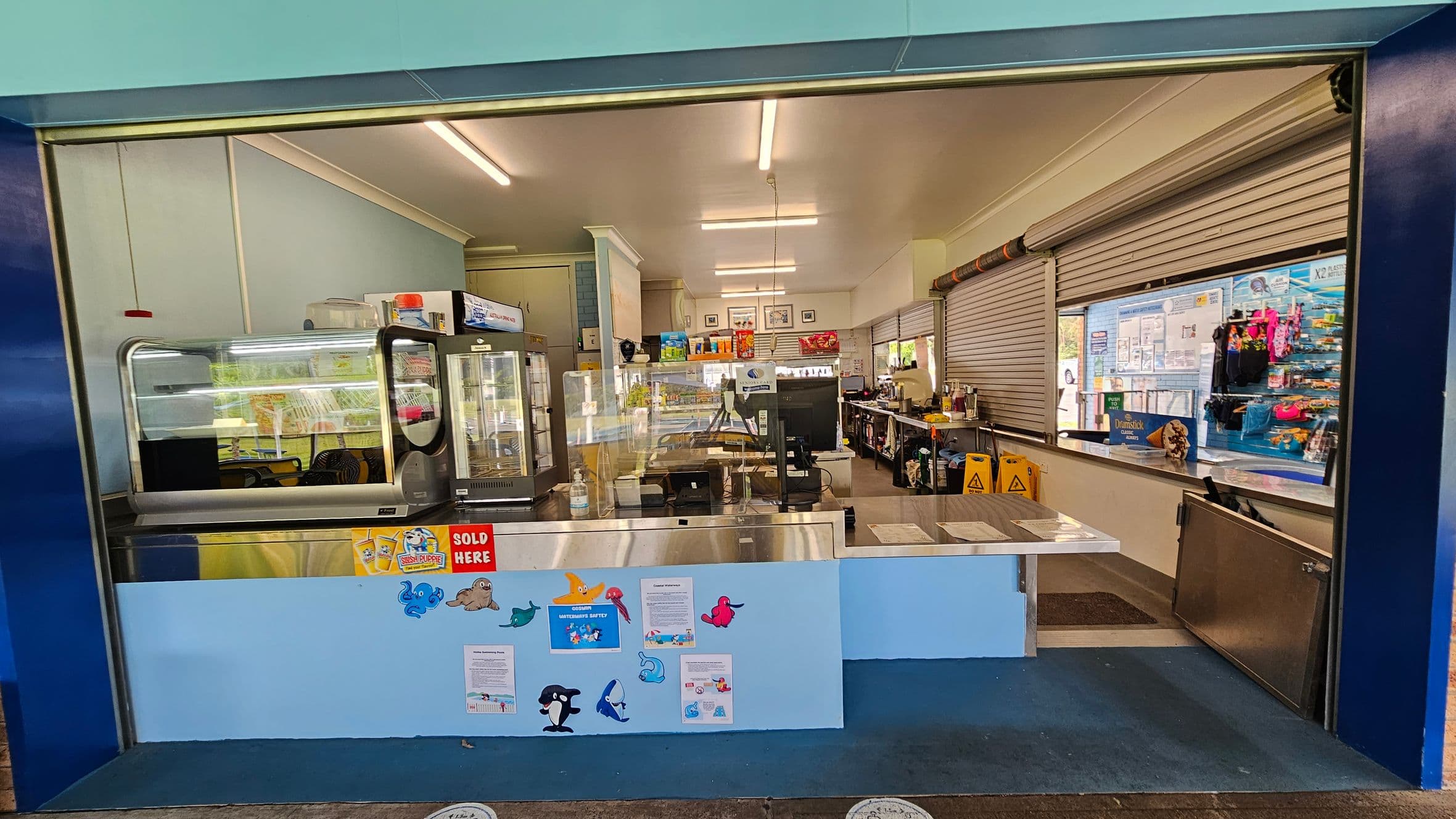 A canteen with a wide serving counter in the foreground, decorated with colourful sea creature cutouts and signs. Behind the counter, there are display fridges and shelves stocked with various items. The interior is well-lit with fluorescent lights, and the walls are light blue. On the right, there are closed roller shutters and a display of snacks and drinks. The floor is blue, and there are caution signs visible inside.