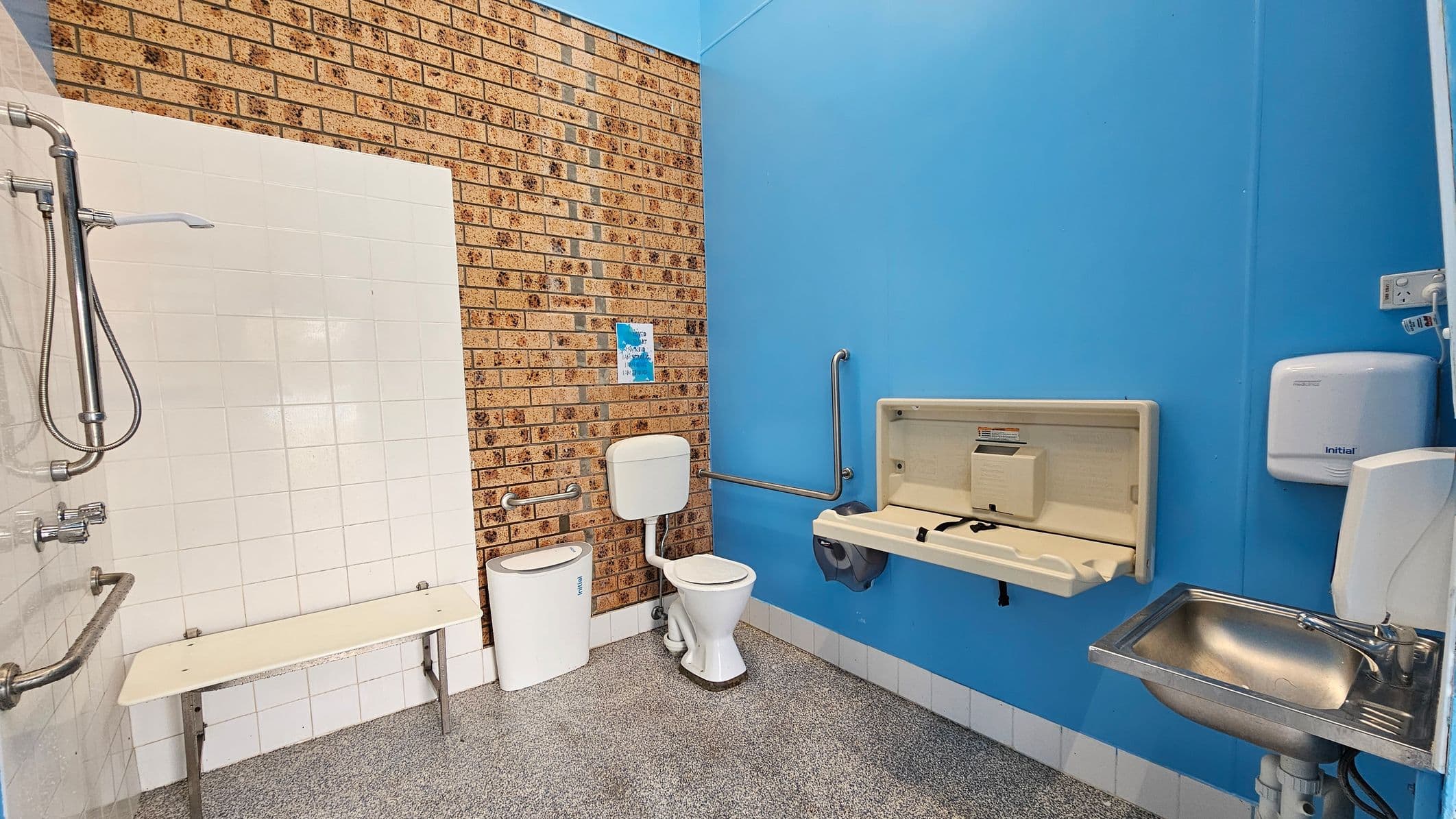 A bathroom with a brick wall on the left and a blue wall on the right. The left side features a shower with a metal handle and a white tiled wall, along with a foldable bench. In the centre, there's a toilet with a white cistern and a small bin beside it. The right side has a baby changing station, a stainless steel sink, and a wall-mounted hand dryer. The floor is speckled grey, and there are grab bars near the toilet and changing station.