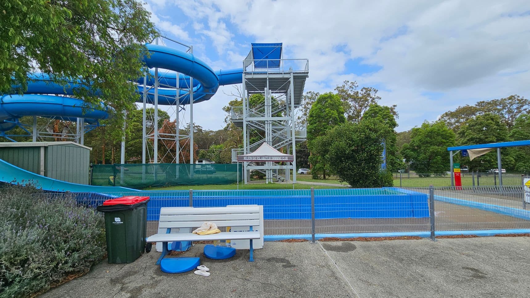 A water park scene with a large blue water slide on the left, supported by a metal structure. In the centre, there's a tall platform with stairs leading up to the slide. A sign reads "Nelson Bay Swimming Club" under a small canopy. In the foreground, a white bench with a towel and flip-flops is next to a green bin with a red lid. The area is fenced, and there are trees and grass in the background. A small shelter with a shade cloth is on the right.