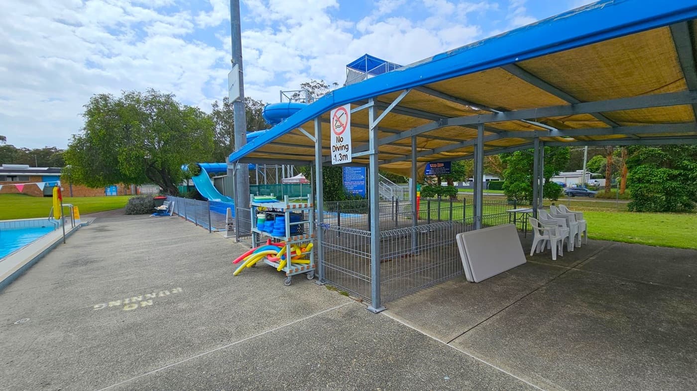 A poolside area with a blue water slide on the left, surrounded by a fence. A paved walkway leads to a covered seating area on the right, with stacked white plastic chairs and a folded table. A sign reads "No Diving 1.3m". Pool equipment, including colourful noodles, is stored on a rack. In the background, there are trees and a grassy area. The ceiling of the covered area is made of a beige material supported by a metal frame.