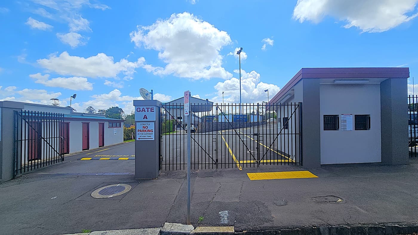 A gated entrance labelled "Gate A" with a sign stating "Strictly No Parking in the Gateway at Any Time" is in the centre. The gate is flanked by a small building on the right with two small windows and a notice. On the left, there are several storage units with brown doors. The ground is paved with yellow markings, and a street sign is visible. In the background, there are tall floodlights and a partially visible structure. The sky is blue with scattered clouds.
