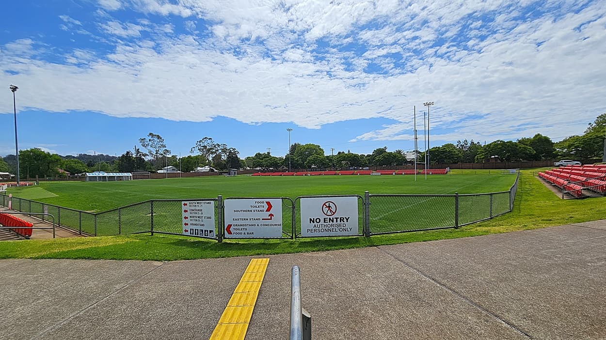 A sports field with a green grass pitch is surrounded by a chain-link fence. In the foreground, a concrete path leads to a gate with signs indicating "No Entry" and directions to various stands and facilities. On the right, there are rows of red stadium seats. Trees and a few buildings are visible in the background, along with tall floodlights. The sky is partly cloudy.