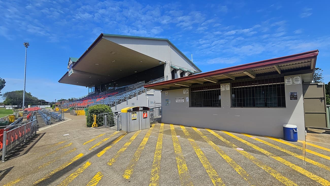 On the left, there is a large grandstand with rows of red and blue seats, partially covered by a sloping roof. A concrete pathway runs alongside, bordered by metal railings and a green hedge. On the right, a wide ramp with metal railings leads upwards, flanked by a small building with a red roof. The foreground features a concrete floor with yellow striped markings, while the ceiling of the grandstand is visible, with several lights attached.