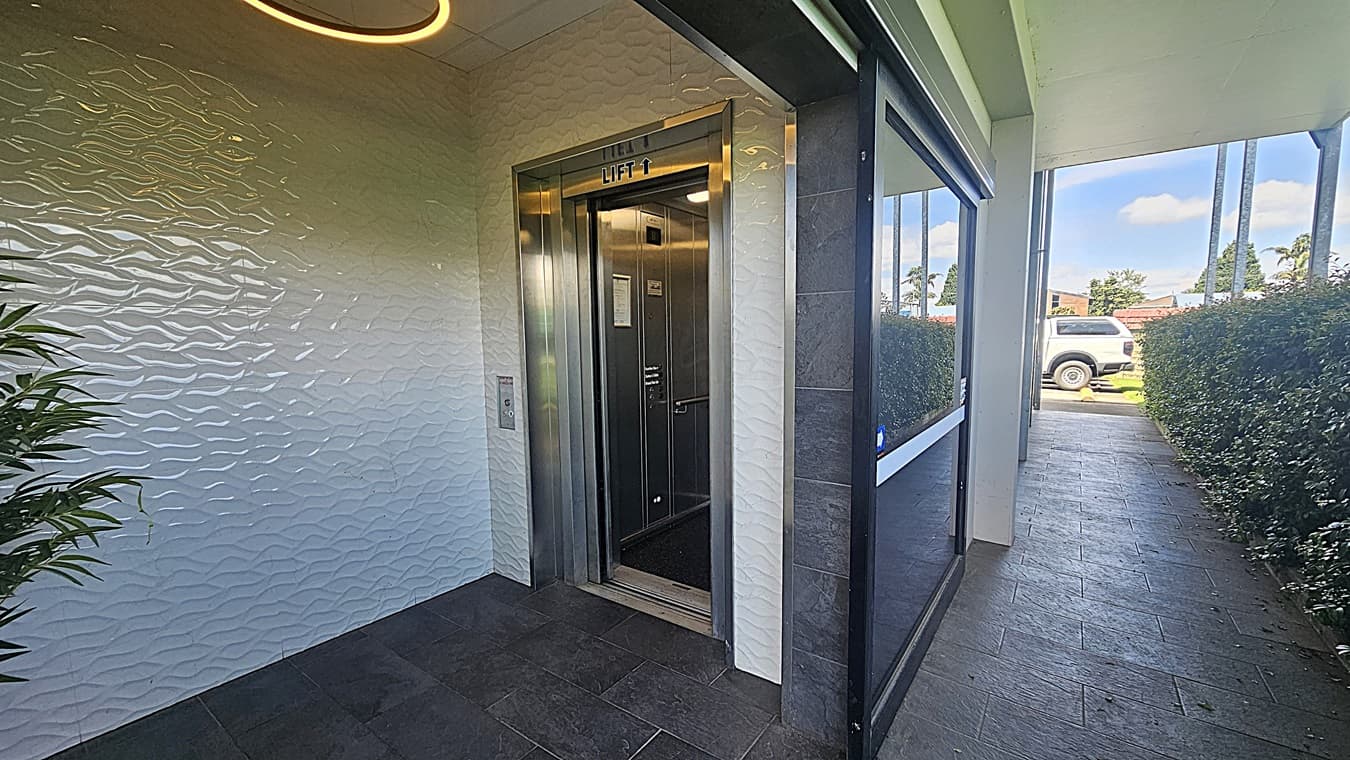 On the left, a textured white wall with a leafy plant in front. An open lift with a metallic frame is visible, labelled "Lift 1". On the right, a glass wall reflects the surroundings, leading to a corridor lined with hedges. The floor is dark stone tiles, and the ceiling has a circular light fixture. In the background, a white vehicle is parked near more greenery.