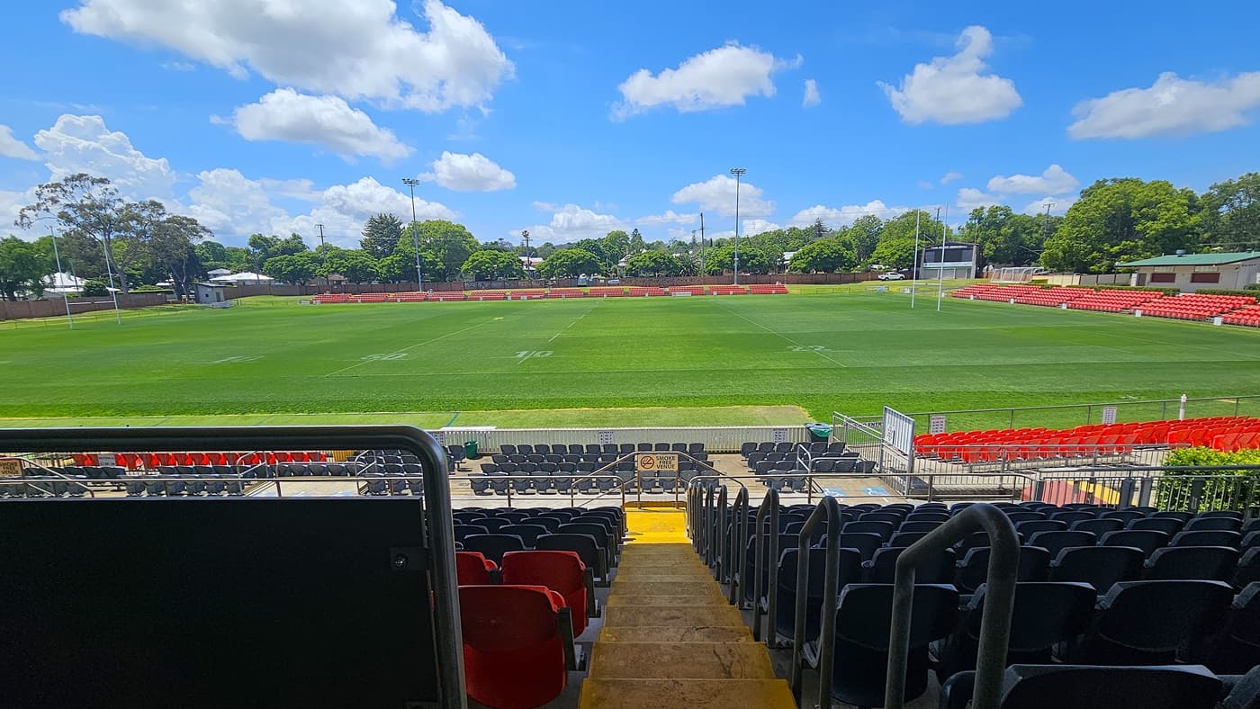 On the left, rows of red seats line the edge of a green sports field. On the right, more red seating continues along the field's perimeter. In the middle, a wide staircase with yellow steps descends between black seats. The background features trees and a small building. The floor is concrete, and the ceiling is not visible.