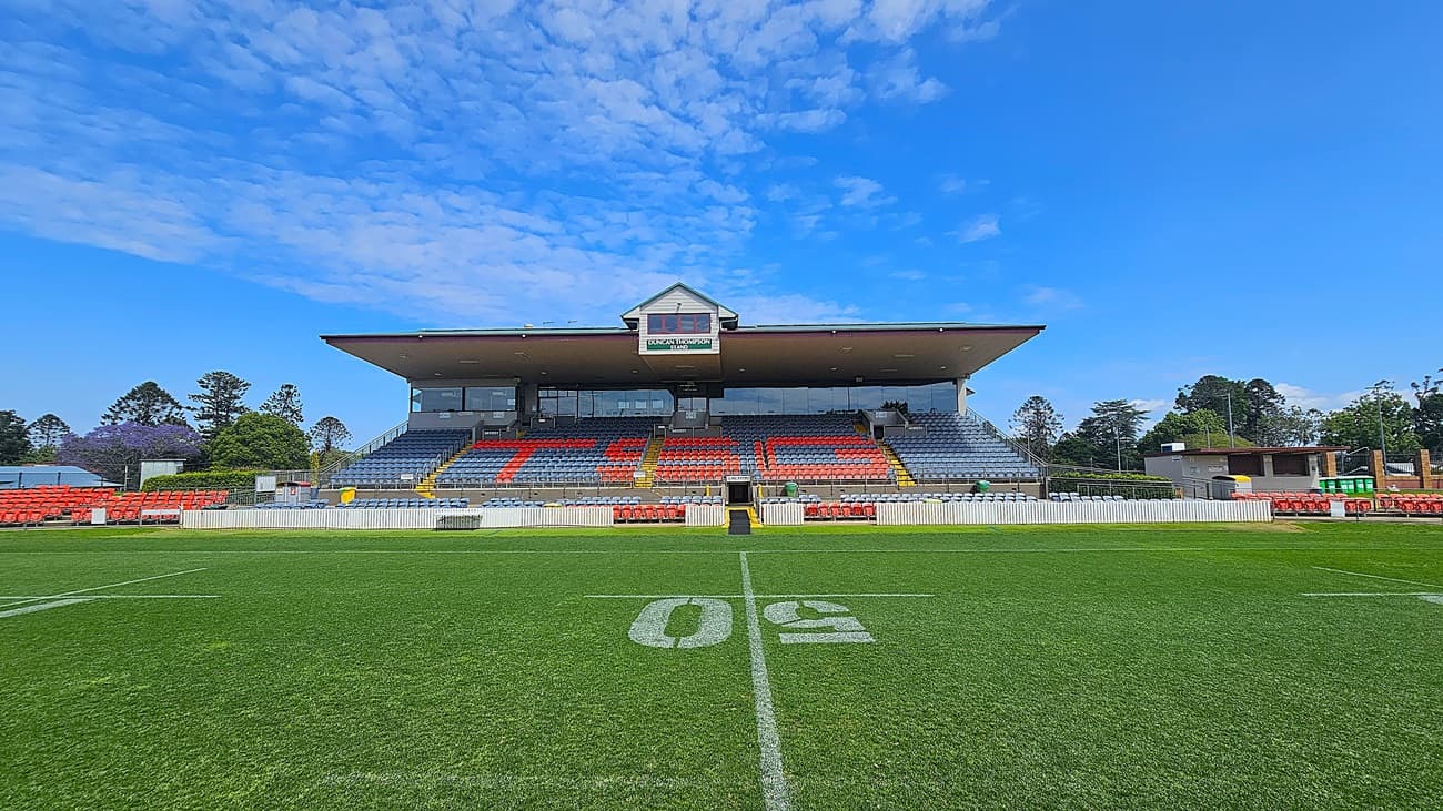 A sports stadium with a large grandstand in the centre, featuring rows of red and blue seats. The letters "TIG" are visible in the seating arrangement. The field in the foreground is marked with a "50" yard line. On the left and right, there are additional seating areas and small buildings. Trees and greenery are visible in the background.