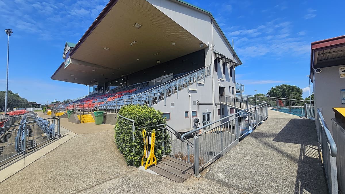 A sports stadium with a large covered grandstand on the left, featuring rows of red and blue seats. A concrete pathway with metal railings leads up to the seating area. A green bush and a yellow barrier are near the entrance. On the right, a smaller building with a corrugated roof is visible. The background includes trees and a clear blue sky.