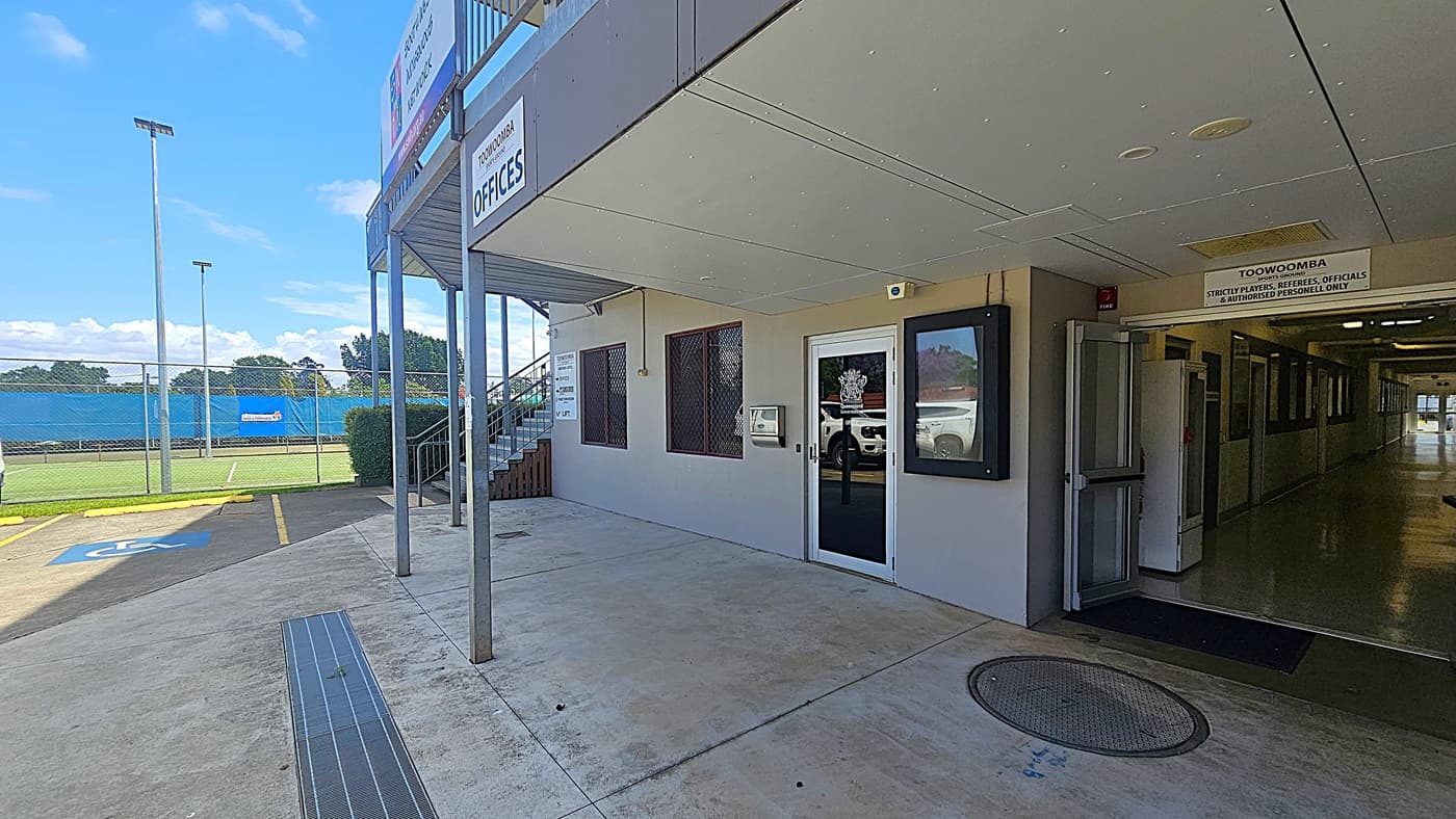 A building exterior with a sign reading "Toowoomba Offices" above the entrance. The entrance has a glass door with a logo and a sign indicating restricted access. To the left, there is a fenced sports field with a blue banner and tall floodlights. The ground is concrete with a marked disabled parking space. A staircase leads to an upper level. The interior hallway is visible through an open door on the right.