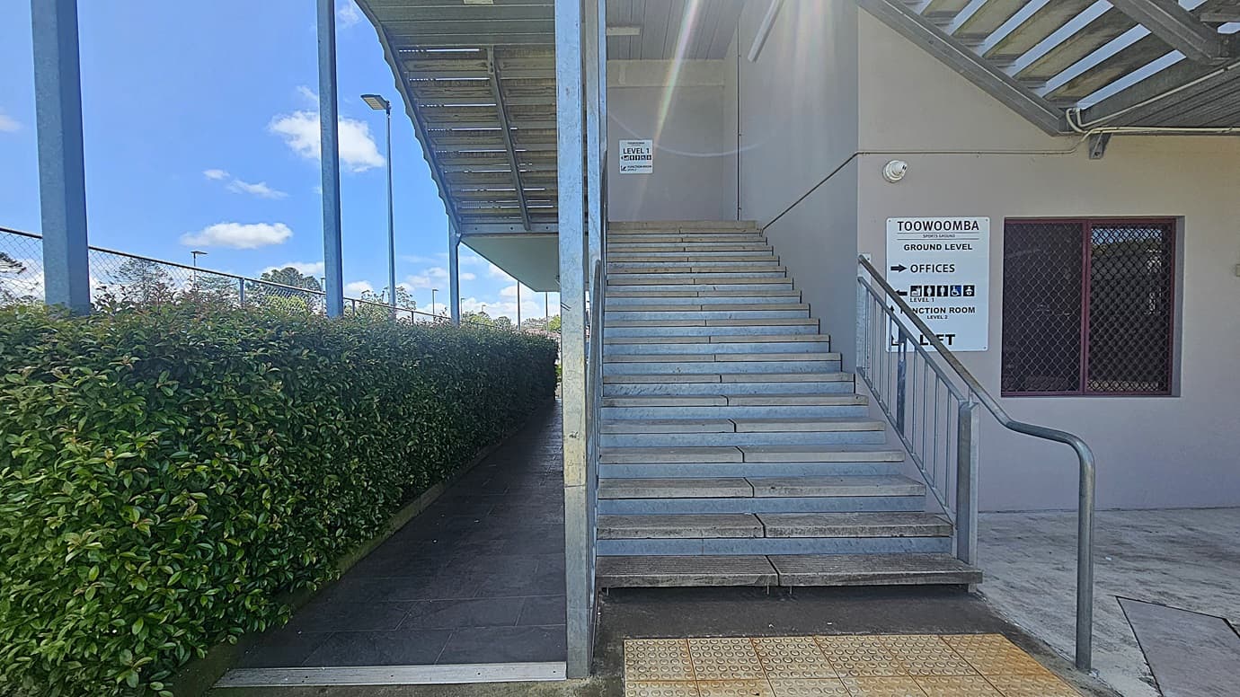 On the left, there is a pathway lined with a tall green hedge and a chain-link fence. On the right, a set of concrete stairs with a metal railing leads up to a building. A sign on the wall indicates directions to various levels and facilities, including offices and a function room. A window with a metal grille is next to the sign. The ceiling above the stairs is covered with metal beams. The floor is tiled with a textured section at the base of the stairs.