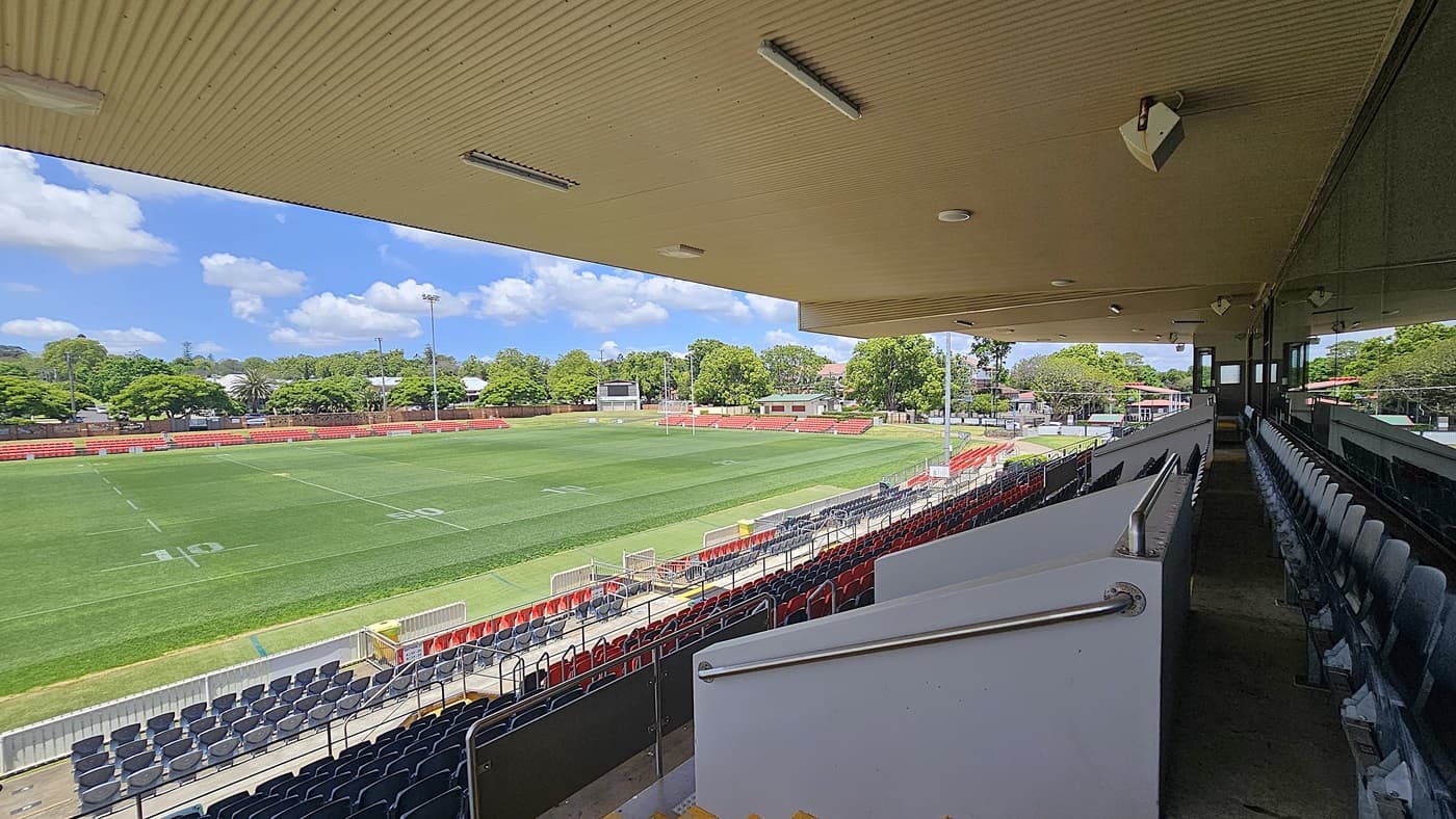 A sports stadium with a green field marked with white lines, surrounded by red and black seating. On the right, there are covered stands with rows of empty seats and a railing. The background features trees and buildings. The ceiling is beige with lights, and the floor of the stands is concrete with yellow steps.