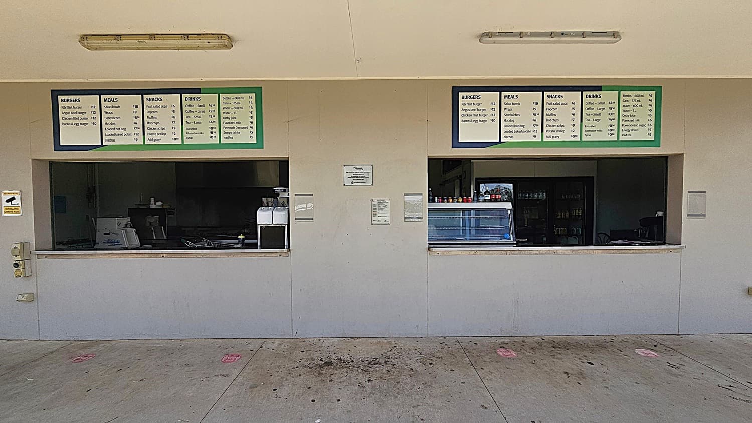 A canteen with two service windows. Above each window are menu boards listing items like burgers, meals, snacks, and drinks. The left window is closed, with a coffee machine visible inside. The right window has a display case and condiment bottles. The wall is light-coloured, and the floor is concrete with some stains. Two fluorescent lights are mounted on the ceiling.