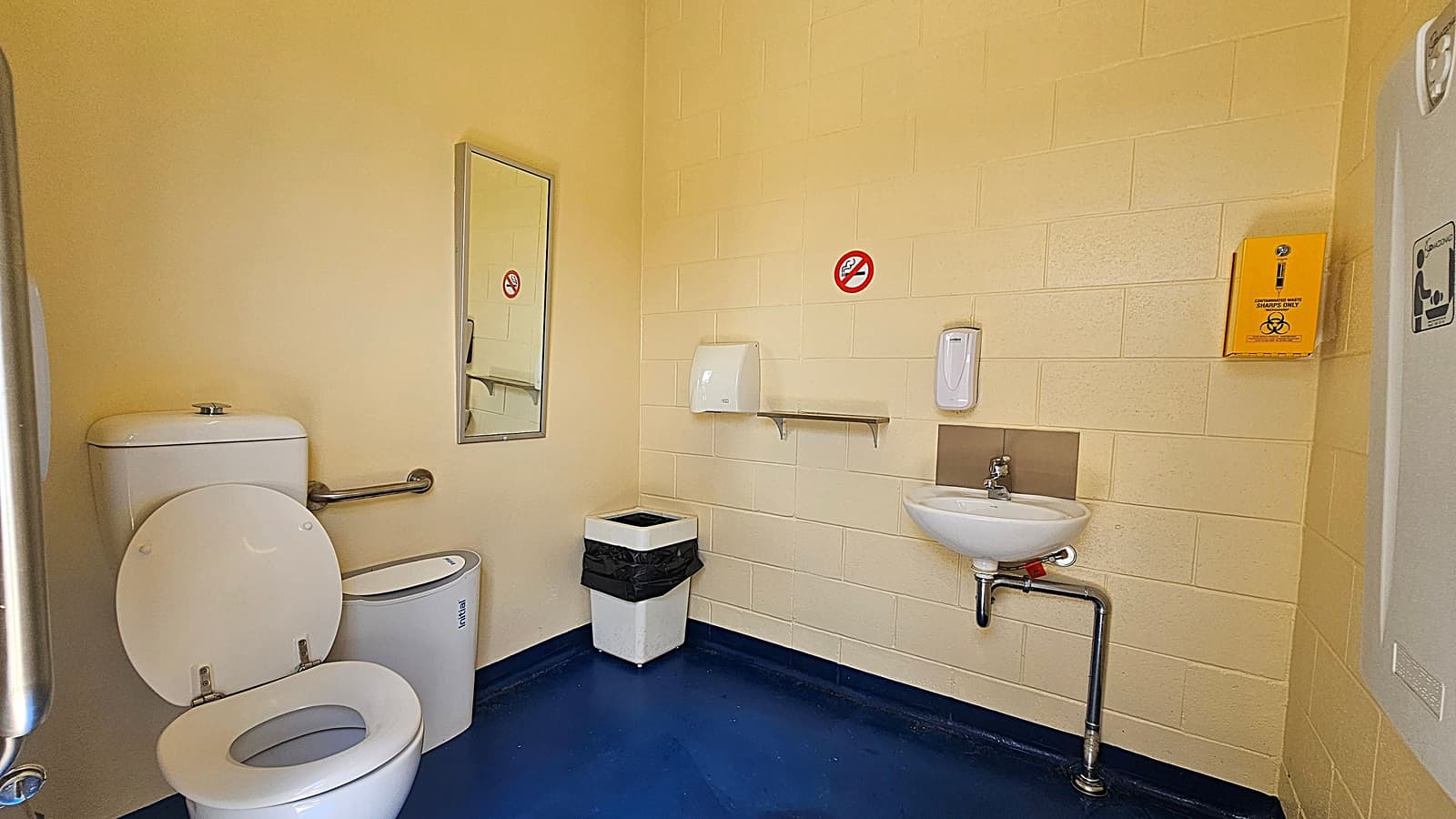 A public toilet with a white toilet and a grab rail on the left. A mirror is mounted on the wall above the toilet. In the centre, there's a bin with a black liner. On the right, a small white sink with a soap dispenser above it. A paper towel dispenser and a no smoking sign are on the wall. A yellow sharps disposal box is mounted on the wall. The floor is dark blue, and the walls are light yellow.