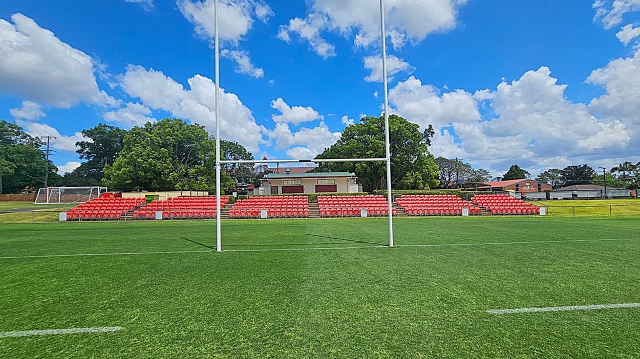 A rugby field with green grass and white goalposts in the foreground. In the background, there are rows of red stadium seats. Behind the seats, there are trees and a building with a beige facade and a red roof. To the left, a football goalpost is visible. The sky is blue with scattered white clouds.