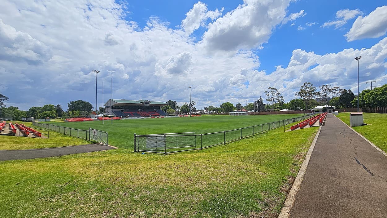A sports field with a well-maintained green pitch is surrounded by a low metal fence. On the left, there are red seating stands, and a larger covered stand is visible in the background. On the right, a paved pathway runs alongside more red seating. Trees and a few buildings are in the distance, and tall floodlights are positioned around the field.