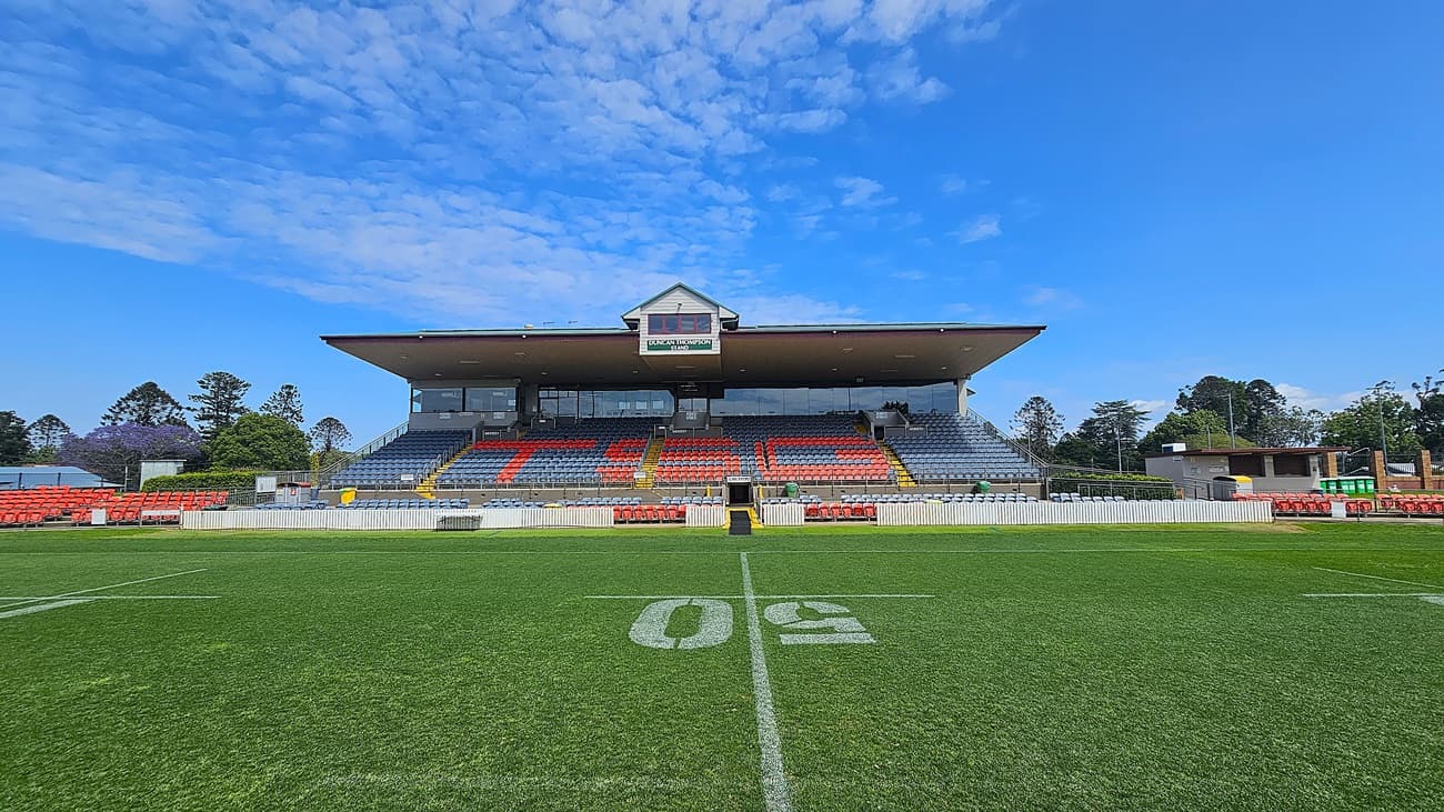 A sports stadium with a green field marked with a large "50" in the centre. The grandstand in the background has rows of red and blue seats, with a small building on top displaying a sign. Trees and additional structures are visible on either side of the grandstand. The sky is clear with some clouds.