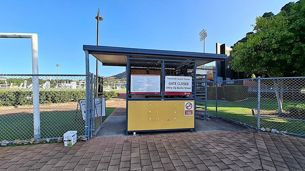 A closed gate at the Townsville Sports Precinct with a sign indicating entry via Burke Street. The gate is part of a metal structure with a yellow panel and a no-smoking sign. To the left, a chain-link fence surrounds a sports field with goalposts. In the background, there are stadium lights and a hill. On the right, a tree and more fencing are visible. The ground is paved with red bricks.
