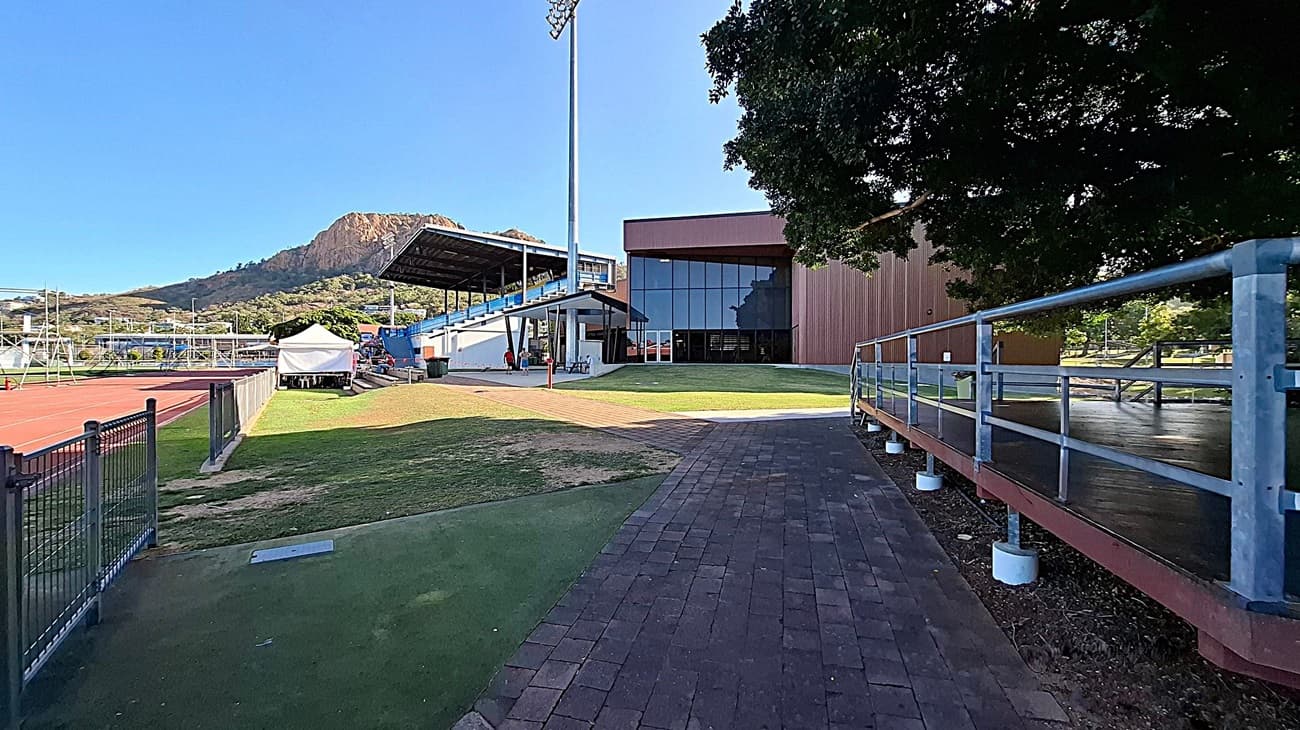 A sports facility with a running track on the left, bordered by a black fence. A white tent is set up near the track. In the centre, there's a building with a large glass facade and a covered seating area. A tall light pole stands nearby. On the right, a tree provides shade over a paved walkway with a metal railing. In the background, a mountain rises, covered in greenery.