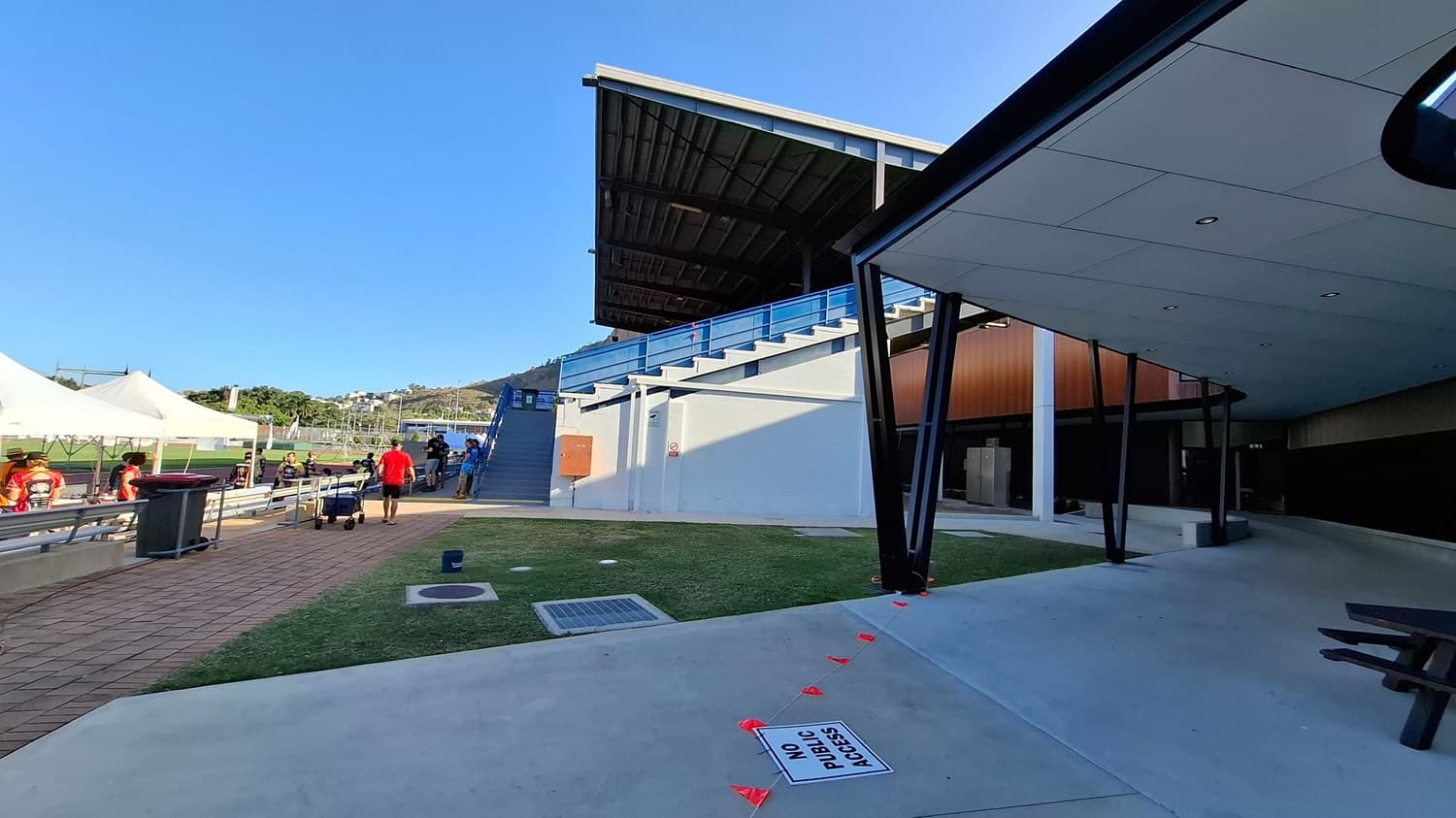 On the left, there are white tents with people sitting underneath, and a pathway with a red bin. In the middle, a man in a red shirt walks along a paved path beside a grassy area. A large grandstand with blue railings is visible, with stairs leading up to it. On the right, a modern structure with a flat roof and black supports is seen, with a "No Public Access" sign on the ground. The ceiling of the structure is smooth with recessed lights.