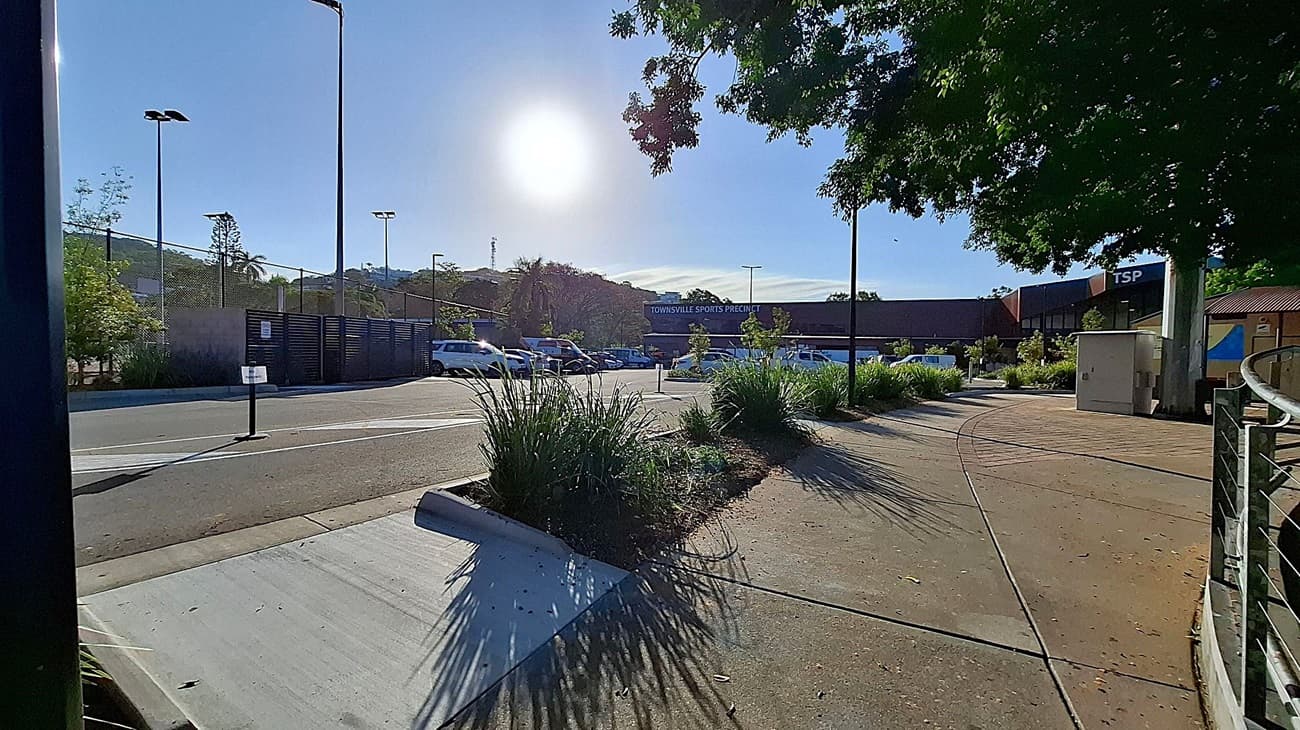 A paved walkway on the right with a metal railing and a utility box. On the left, a road with parked cars and tall streetlights. In the centre, a sign reads "Townsville Sports Precinct" on a building. Greenery, including trees and bushes, is scattered throughout. The sun is bright in the sky, casting shadows on the ground.
