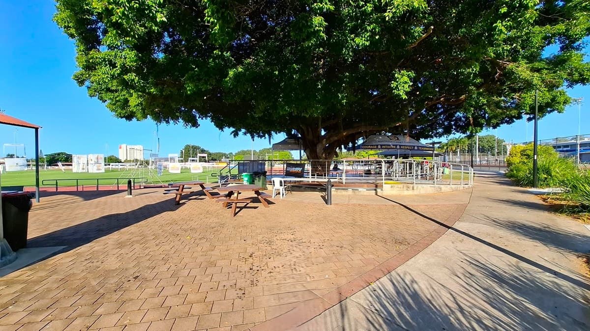A large tree with dense foliage provides shade over a paved area with picnic tables and chairs. To the left, a small structure with a red roof is partially visible. In the background, a sports field with nets and a tall building can be seen. A pathway curves to the right, bordered by grass and plants. A shadow of a person holding a camera is cast on the ground in the foreground.