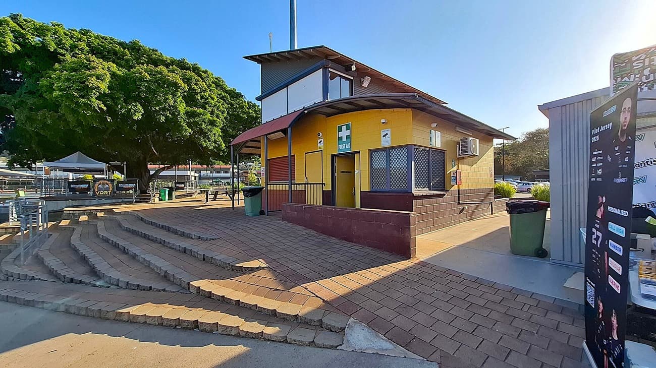 A yellow building with a brown base and a first aid sign is on the right. It has a small awning and barred windows. In front, there are brick steps leading up to a paved area. A large tree with dense foliage is on the left, providing shade. A green bin is near the building, and a poster stand is on the far right. In the background, there are tables, chairs, and a white canopy.