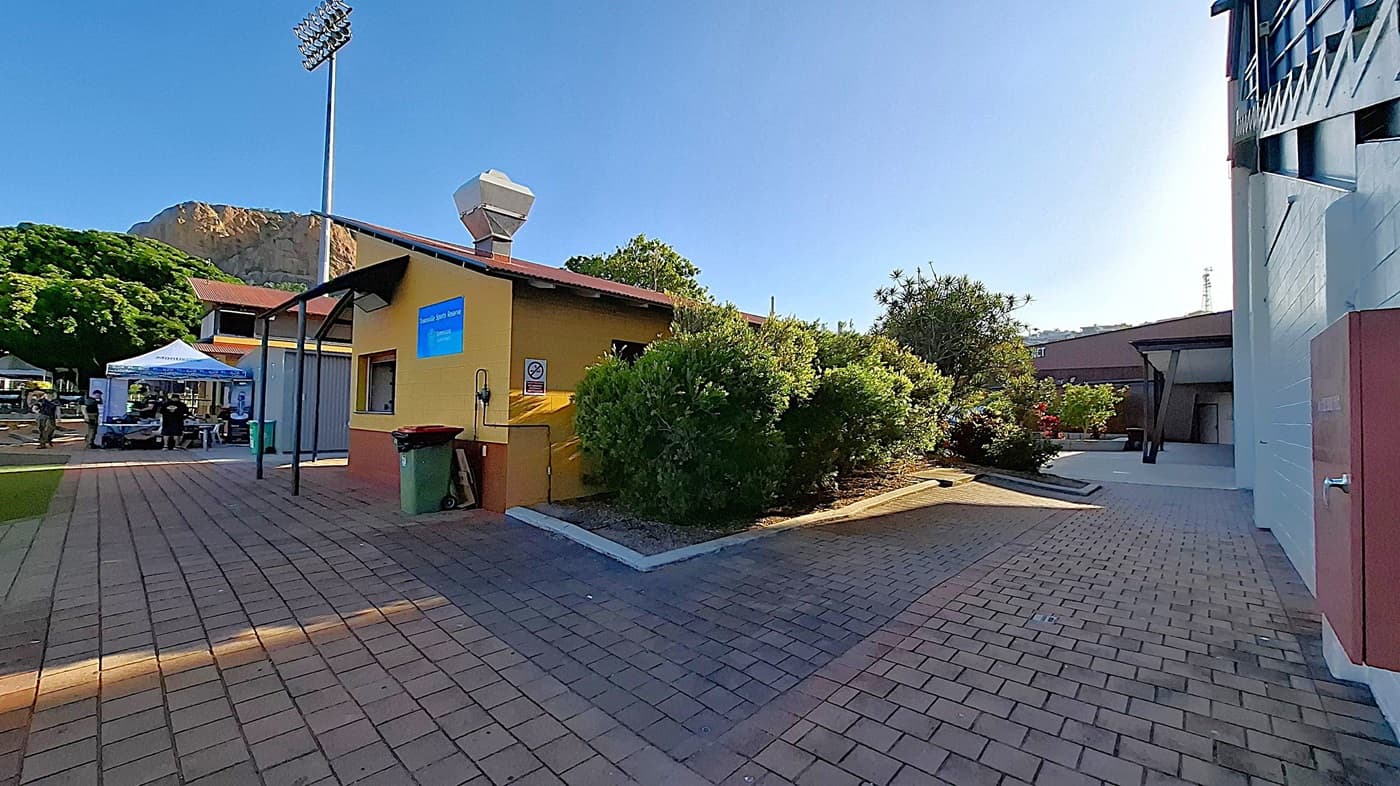 A paved walkway with a yellow building on the left, featuring a sign and a green bin. Bushes and trees surround the building. To the left, a white tent with people underneath is visible. A tall floodlight stands nearby. On the right, a grey building with a covered entrance is partially visible. In the background, a large rocky hill and more greenery can be seen.