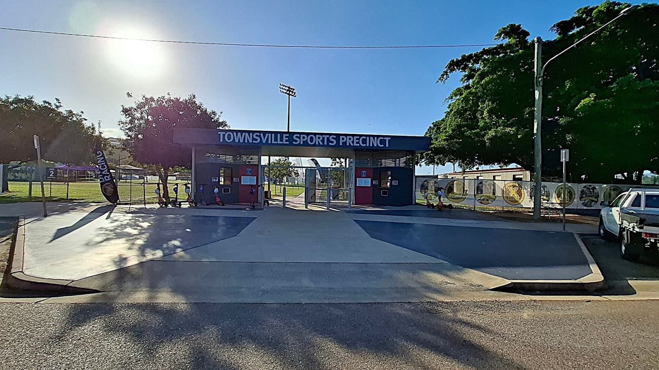 Entrance to the Townsville Sports Precinct with a sign above. In the foreground, a paved area with shadows from nearby trees. On the left, a flag with "RACING" and a fenced grassy area. On the right, a white utility vehicle parked near a fence with circular designs. Trees provide shade on both sides, and a streetlamp is visible on the right.