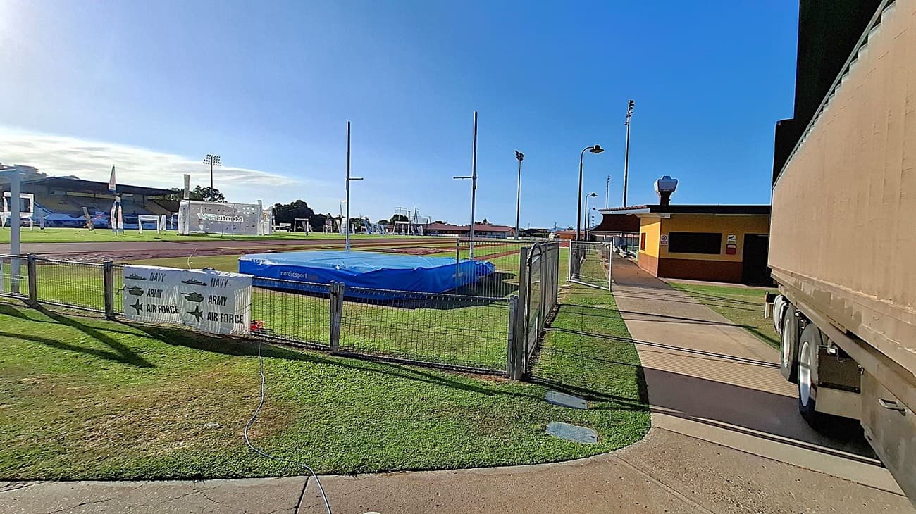 On the left, a sports field with a blue high jump mat and poles is surrounded by a metal fence. Signs on the fence read "Navy," "Army," and "Air Force." In the middle, a paved path runs alongside the field. On the right, a large vehicle is partially visible, parked near a yellow and brown building. Floodlights and a clear blue sky are in the background. The ground is grassy with some concrete areas.